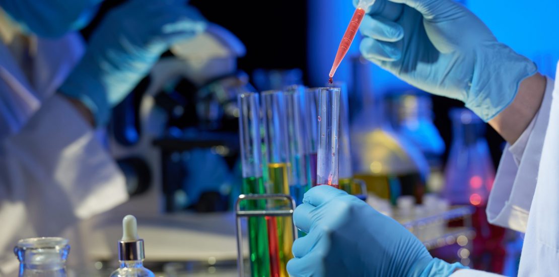 Talented researcher wearing rubber gloves standing at laboratory bench and pouring red liquid into test tube with pipette while working on joint project with colleague