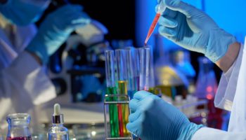 Talented researcher wearing rubber gloves standing at laboratory bench and pouring red liquid into test tube with pipette while working on joint project with colleague